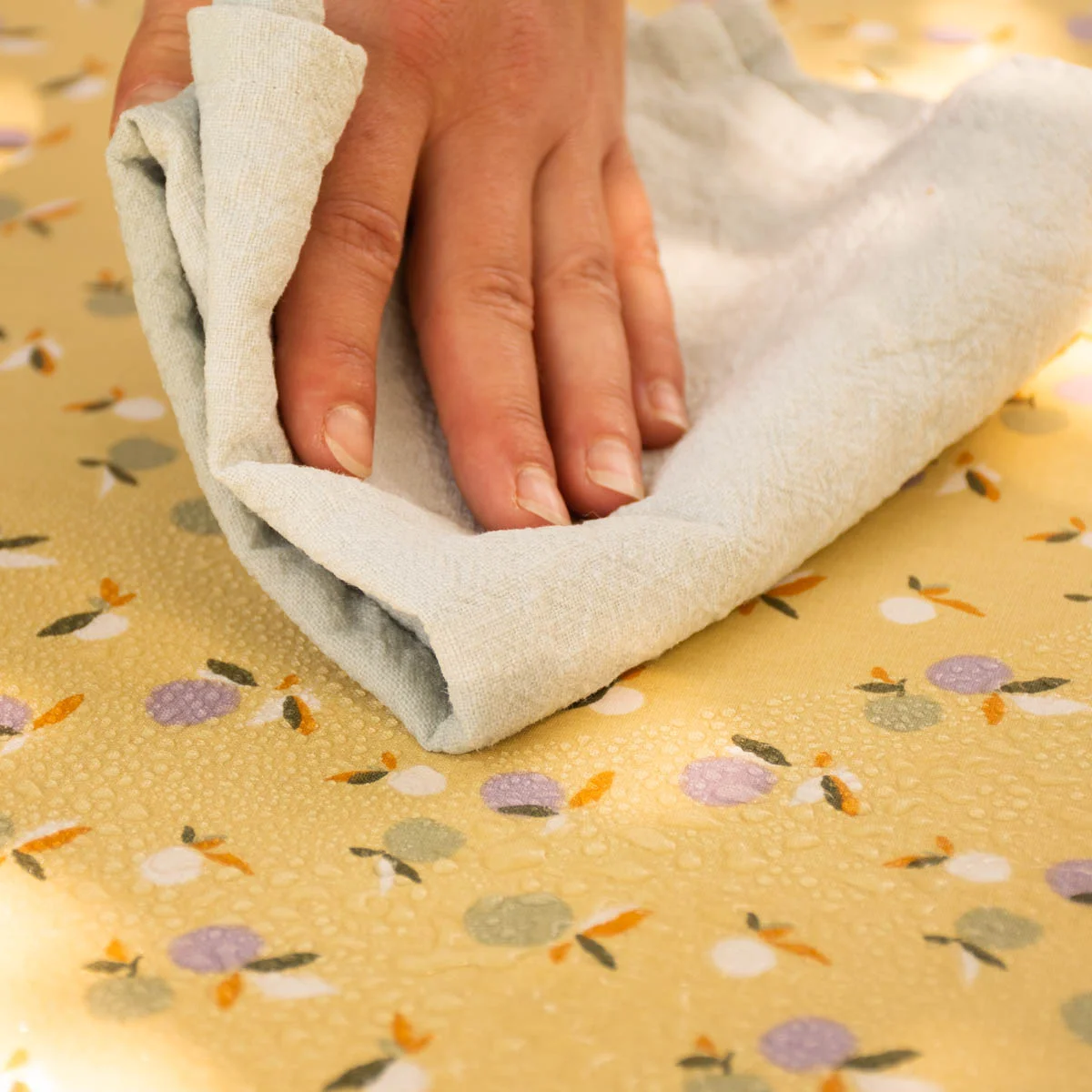 abwaschbare Tischdecke in Hellgelb mit Früchten in weiß, helllila und hellgrün mit orangefarbenen und dunkelgrünen Blättern mit abperlenden Wassertropfen die mit einem trockenen Tuch abgewischt werden.