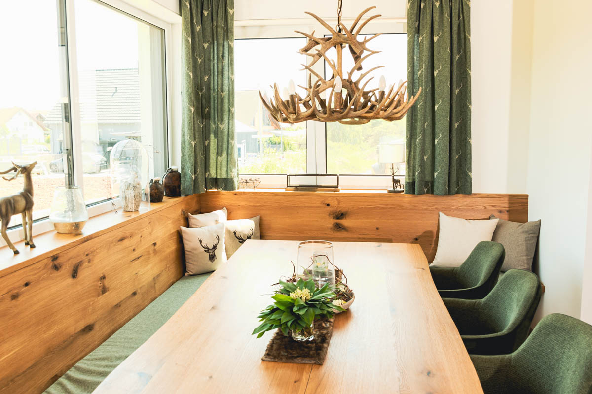 Esszimmer im Landhausstil: Ein Holzesstisch mit einer Holzbank, die eine Bankauflage hat, dahinter farblich passende Vorhänge in grün mit Hirschmotiv