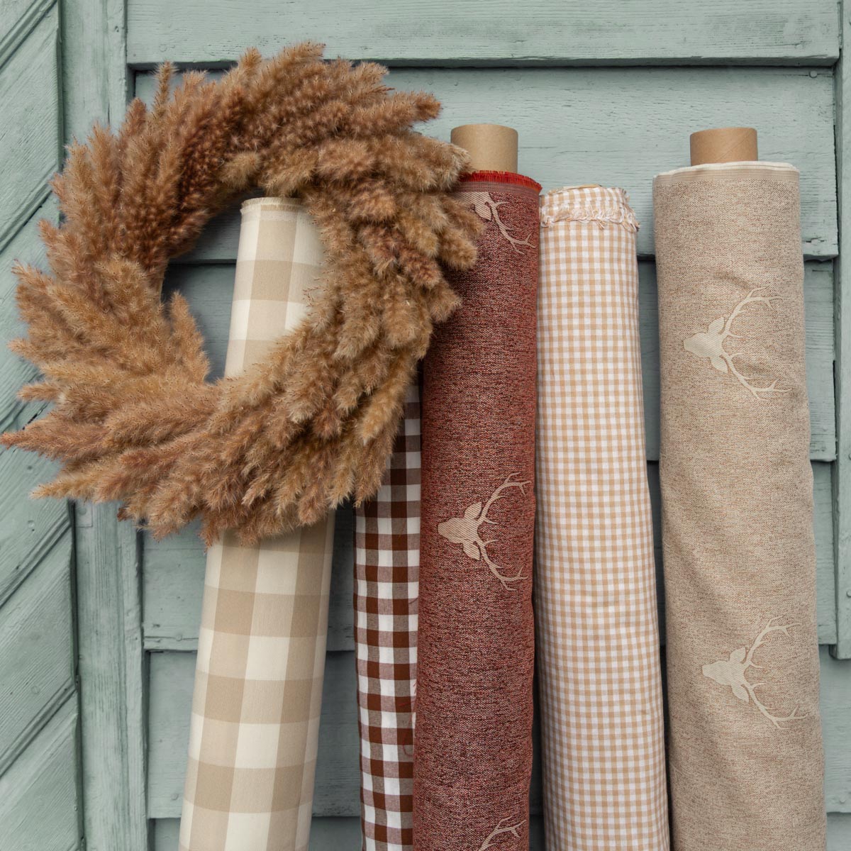 Landhausstoffe im Herbst: Stoffrollen mit Karo und Hirsch-Motiv vor einer grünen Holzwand, dazu ein Pampasgras-Kranz